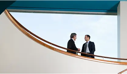Two men standing on a balcony outside a boardroom having a serious conversation